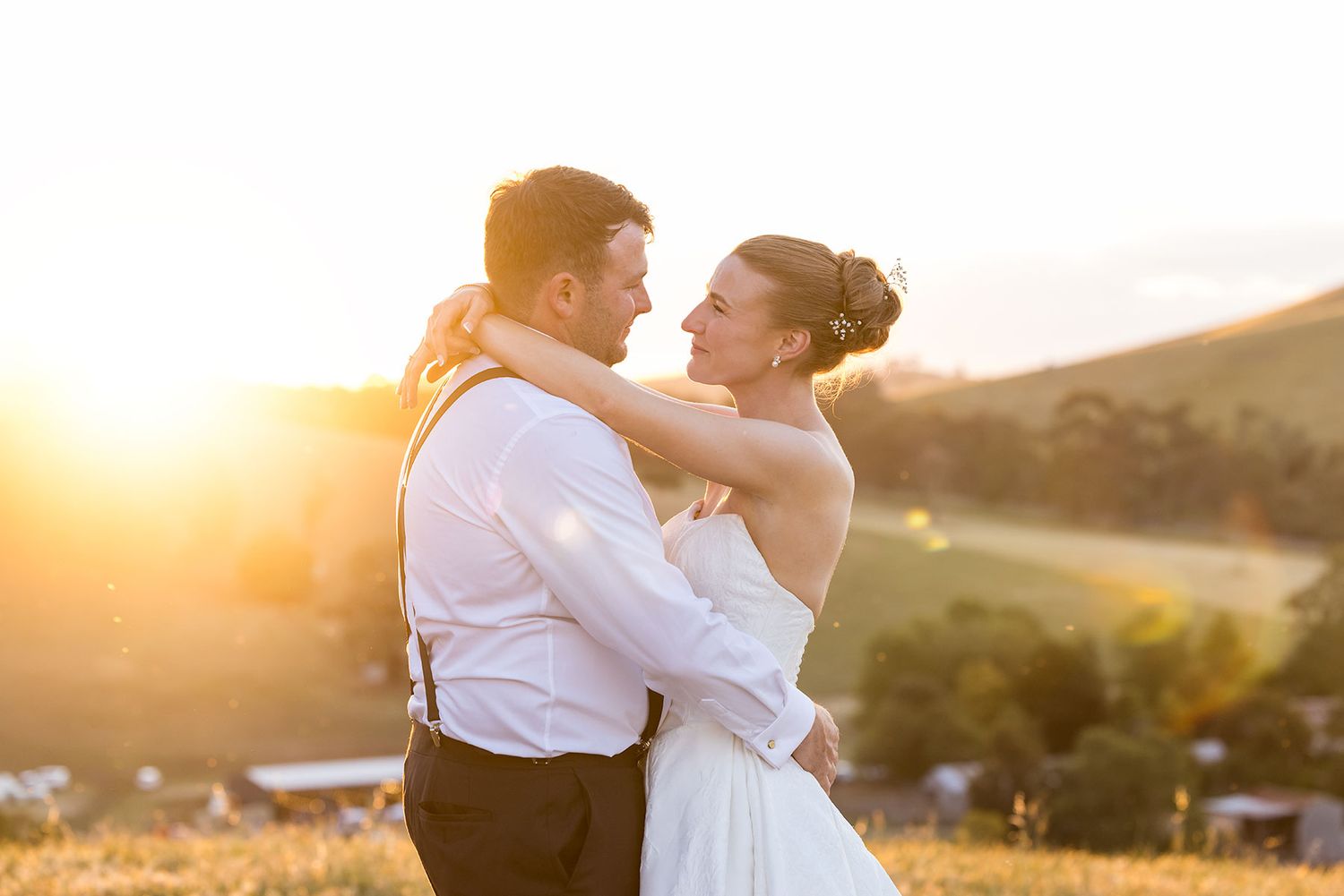 A romantic couple embraces during golden hour sunset in a scenic countryside setting with rolling hills in the background.