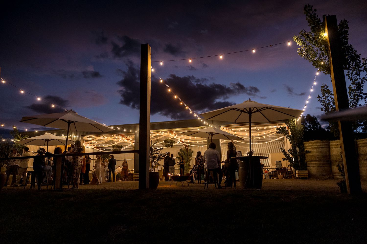 Outdoor evening event space illuminated by string lights with silhouettes of guests beneath umbrellas and tents at a farm wedding.