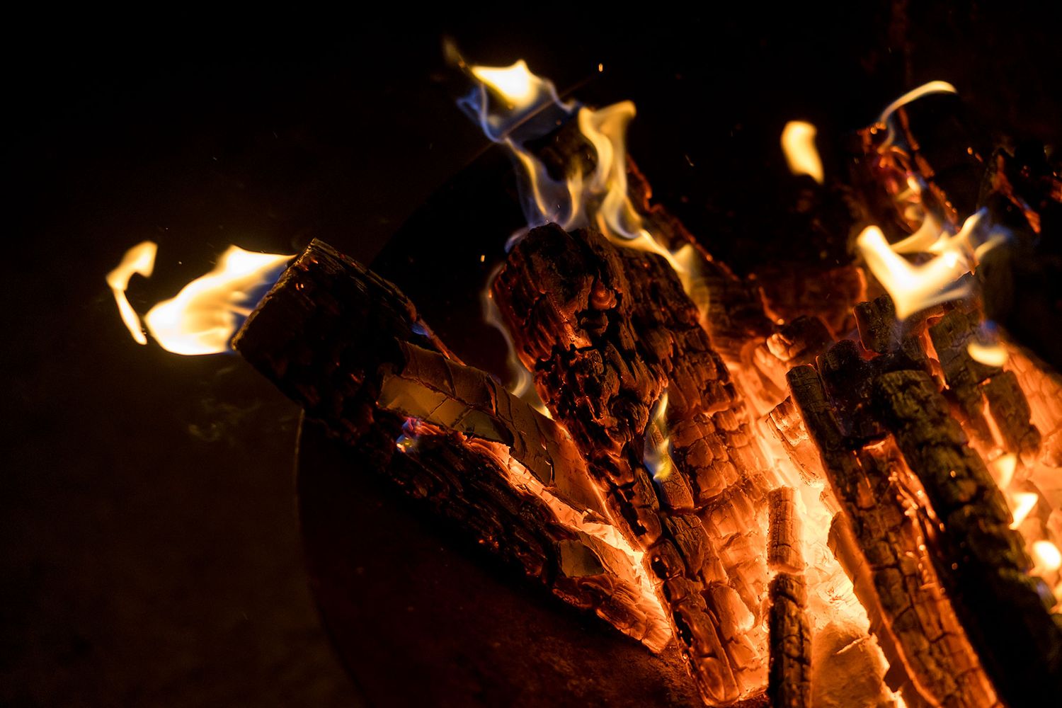 Glowing orange flames dance and flicker on burning logs in a dark nighttime campfire.