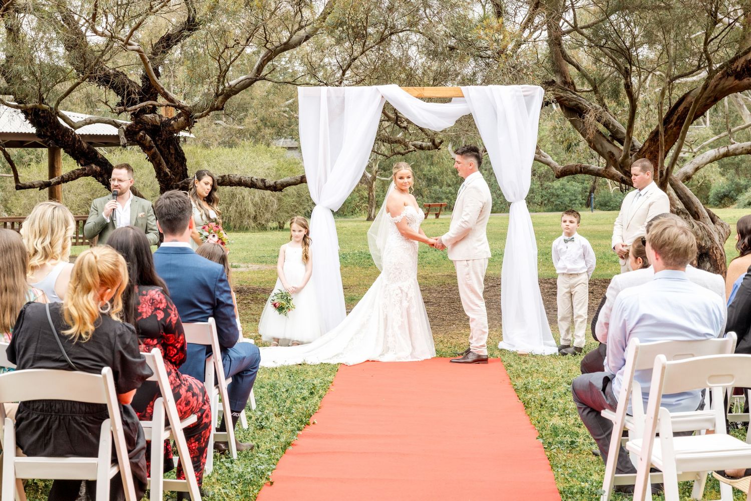 Outdoor wedding ceremony setup with white chairs and floral arch under trees.
