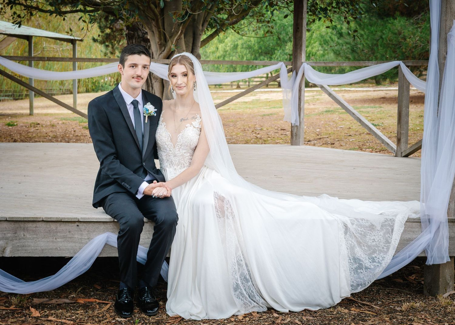 Newlyweds pose together on a wooden bench in an outdoor setting with decorative white draping.