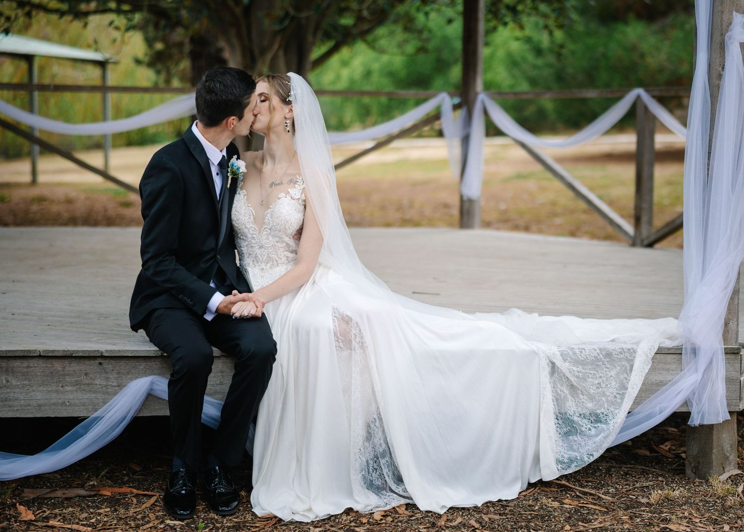 A romantic wedding portrait of newlyweds sitting on a rustic bench with flowing veil.