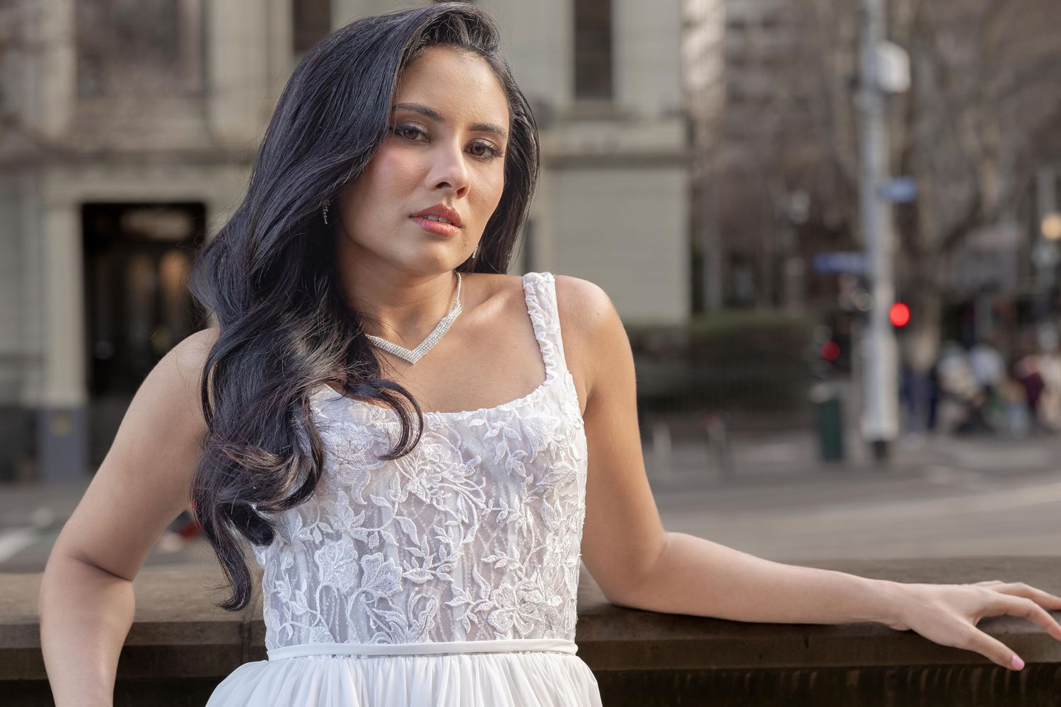 A woman in a white lace dress poses against an urban backdrop.
