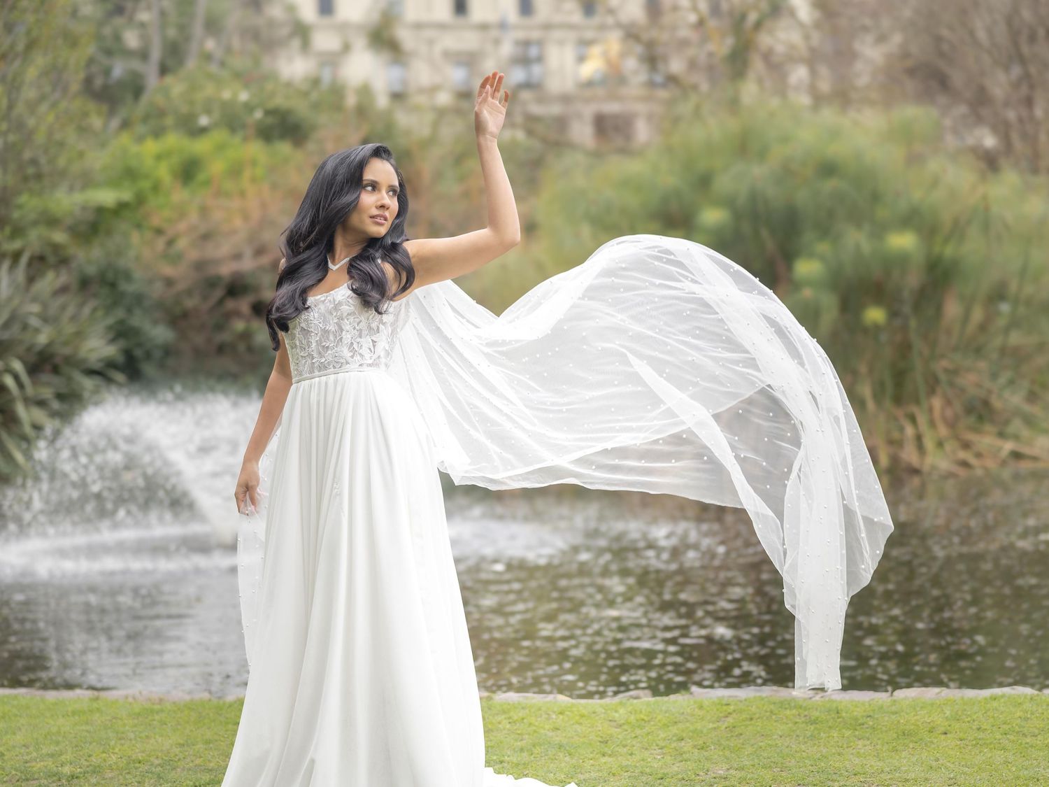 A flowing white wedding dress is showcased against a scenic lake backdrop with classical architecture.