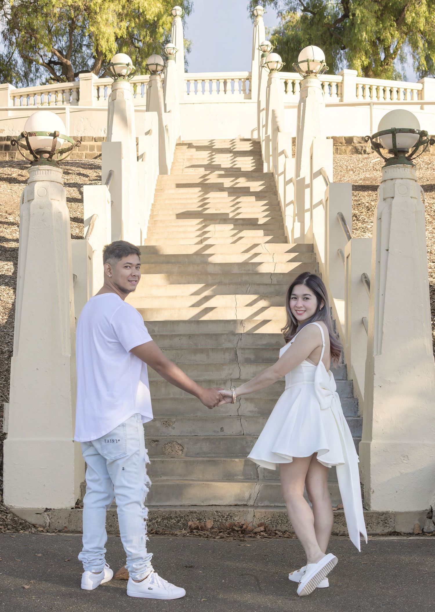 A couple holding hands on white stone steps in matching light-colored outfits.