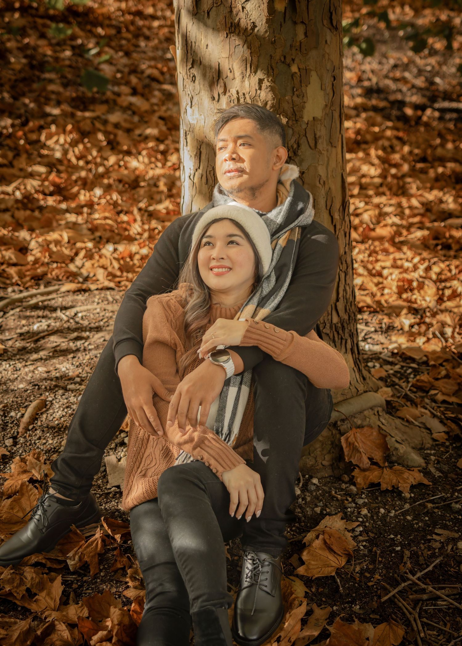 A romantic autumn portrait of a couple sitting against a tree trunk surrounded by fallen leaves.