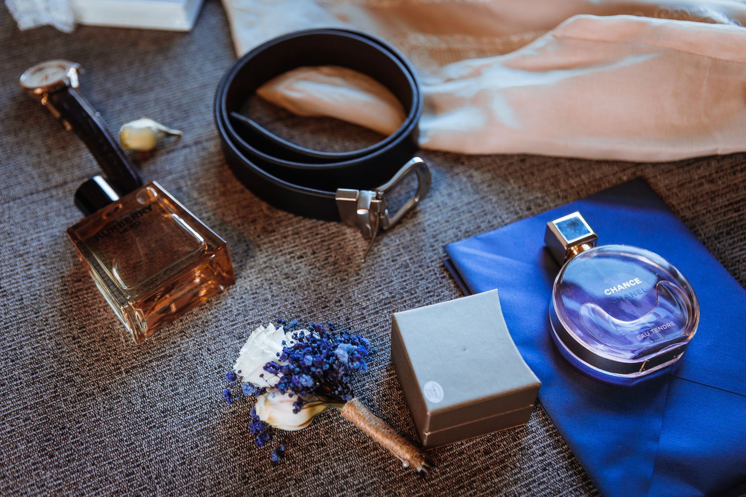 Wedding accessories including perfume, leather belt, and blue boutonniere arranged on dark surface.