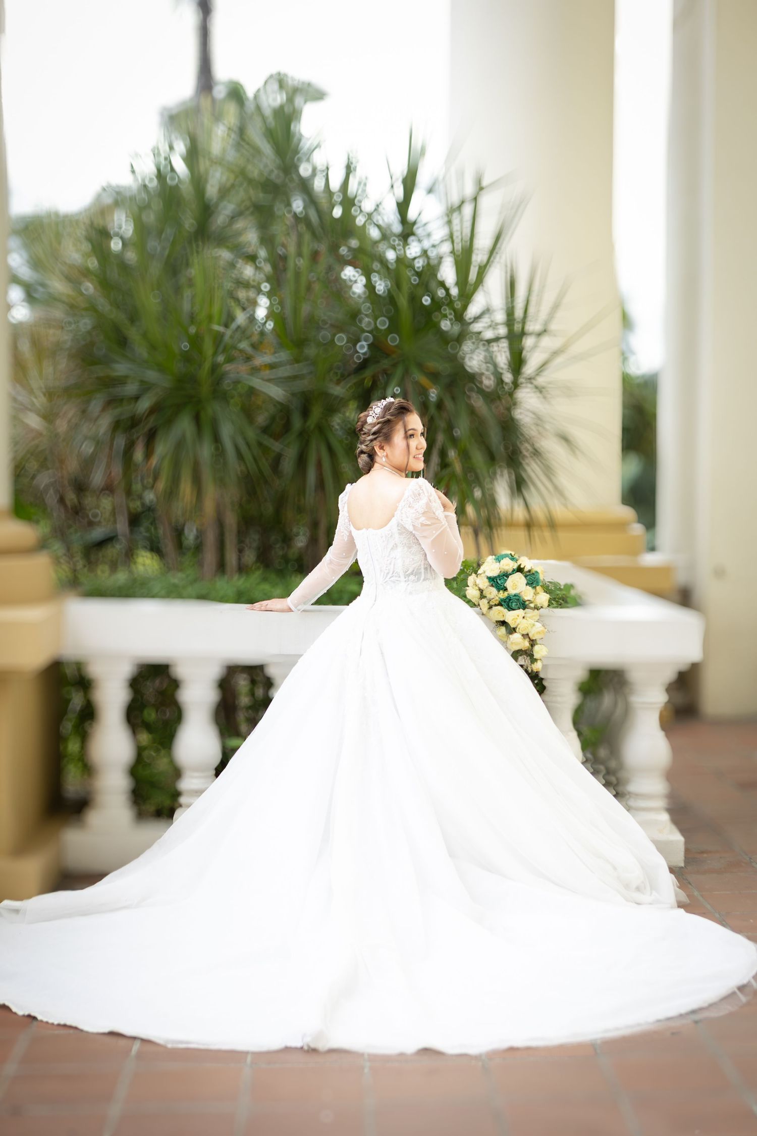 Elegant bride in long-sleeved lace wedding gown with flowing train poses on terrace with tropical plants in background.