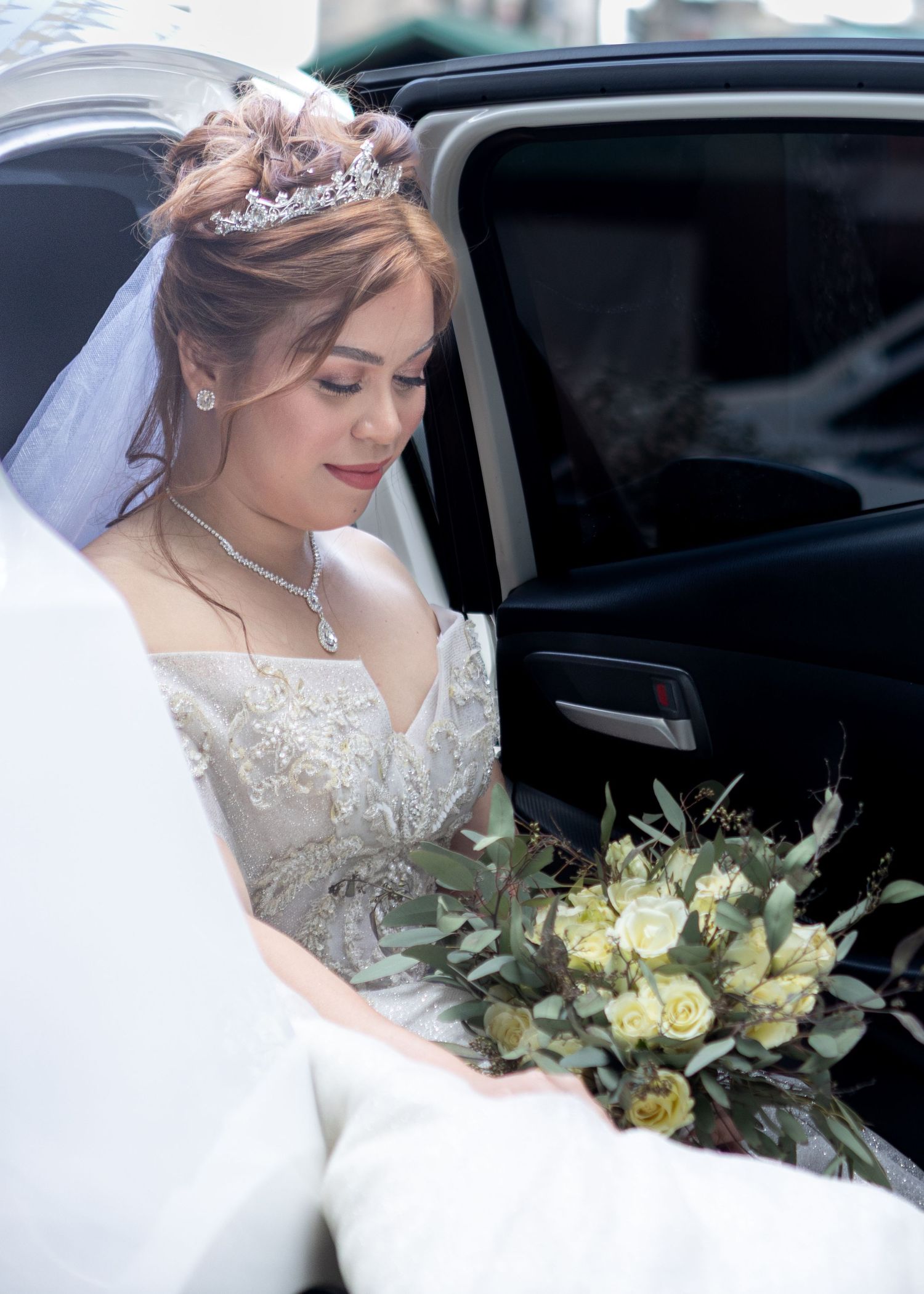 Bride wearing tiara and veil sits in luxury car with white and green bouquet.