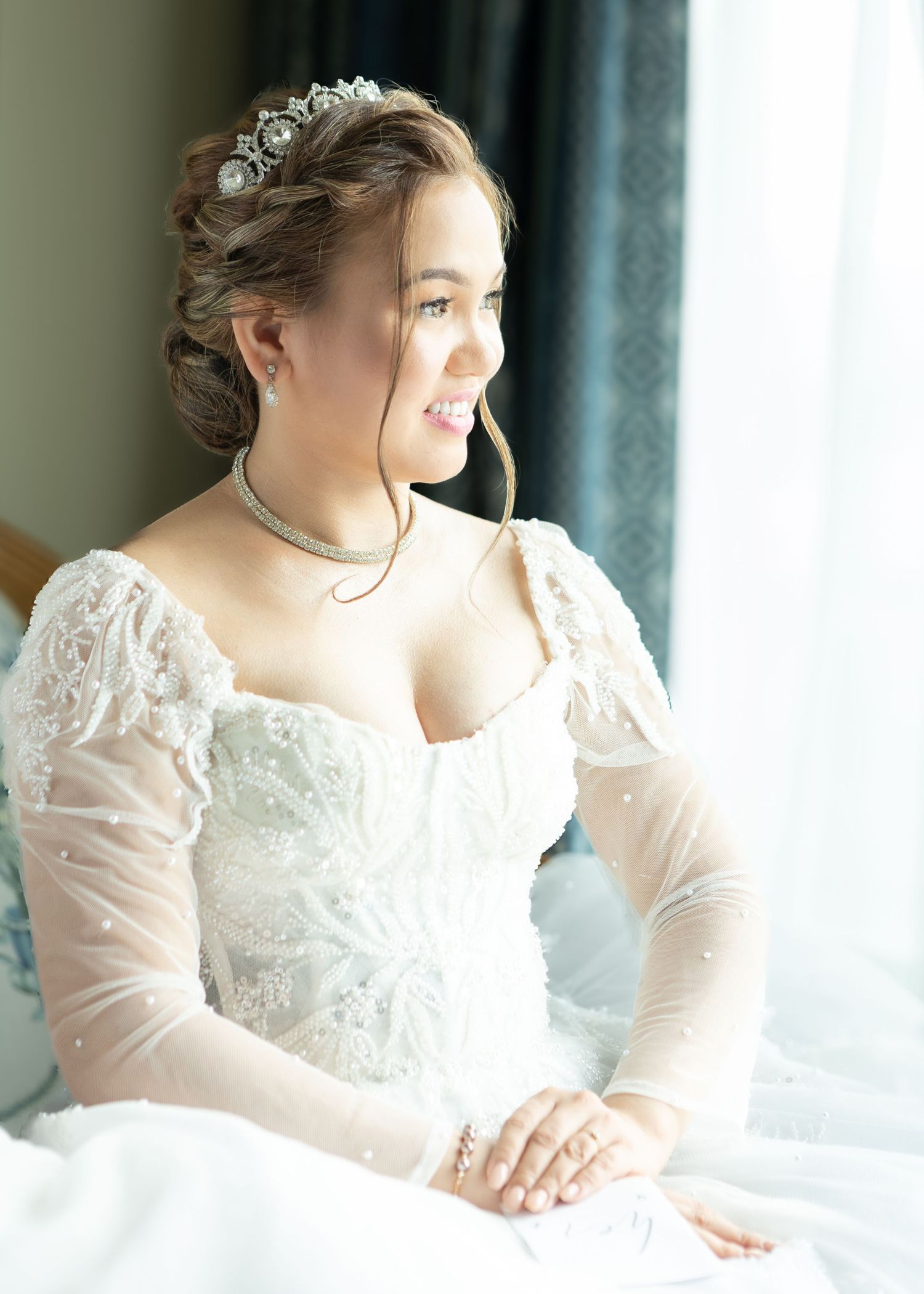 Bride in elegant lace wedding dress with sheer sleeves poses gracefully by window in bridal portrait series.