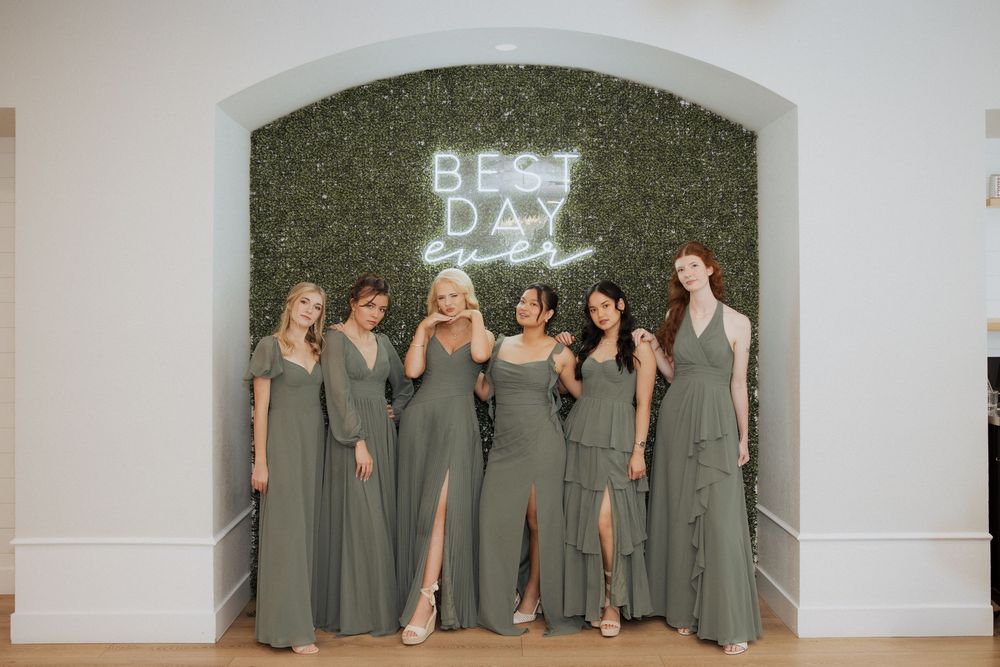 Bridesmaids in sage green floor-length dresses pose together under a neon 'Best Day' sign at a wedding venue.
