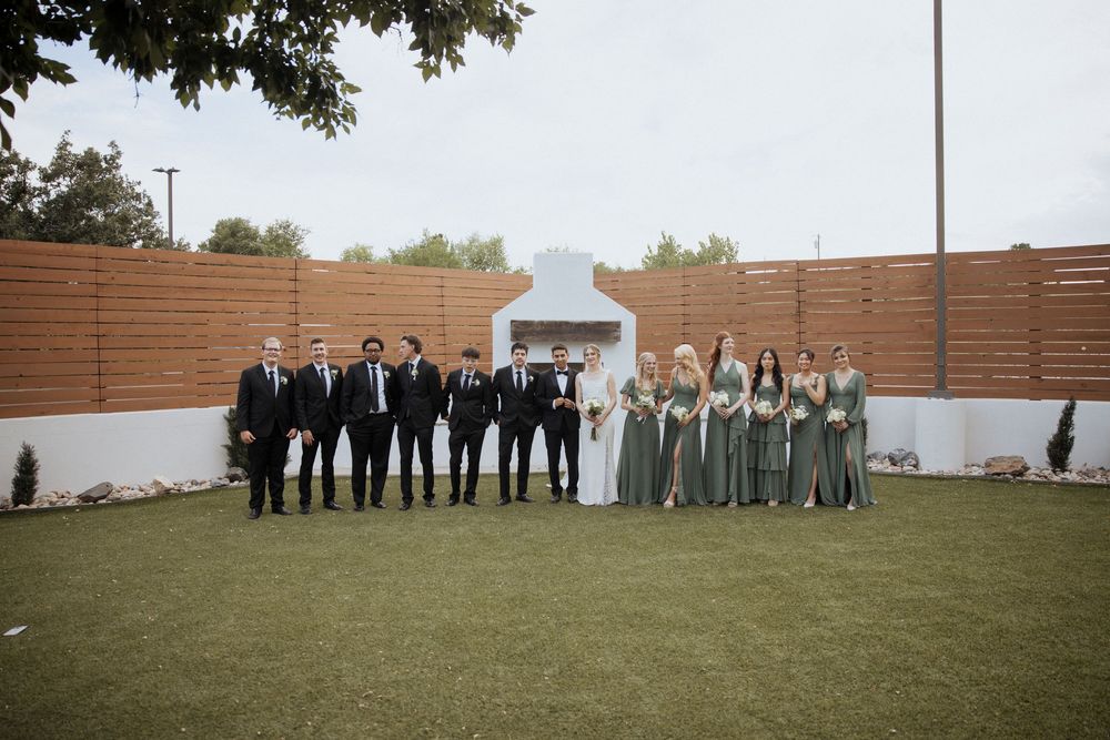 Wedding party members stand together in formal attire against wooden fence and outdoor backdrop for group photos.
