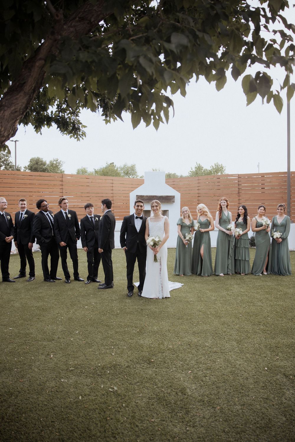 Wedding party stands on green lawn with groomsmen in black suits and bridesmaids in sage green dresses.
