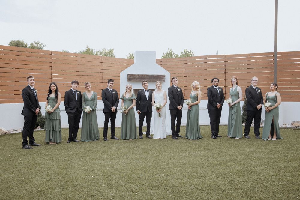 Wedding party members in sage green dresses and dark suits pose together on grass in front of wooden wall backdrop.