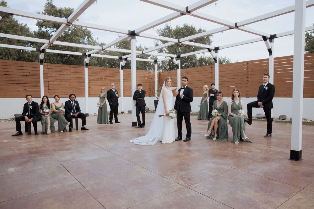 Wedding party arranged in formal poses under a white tent structure with wooden walls in the background.