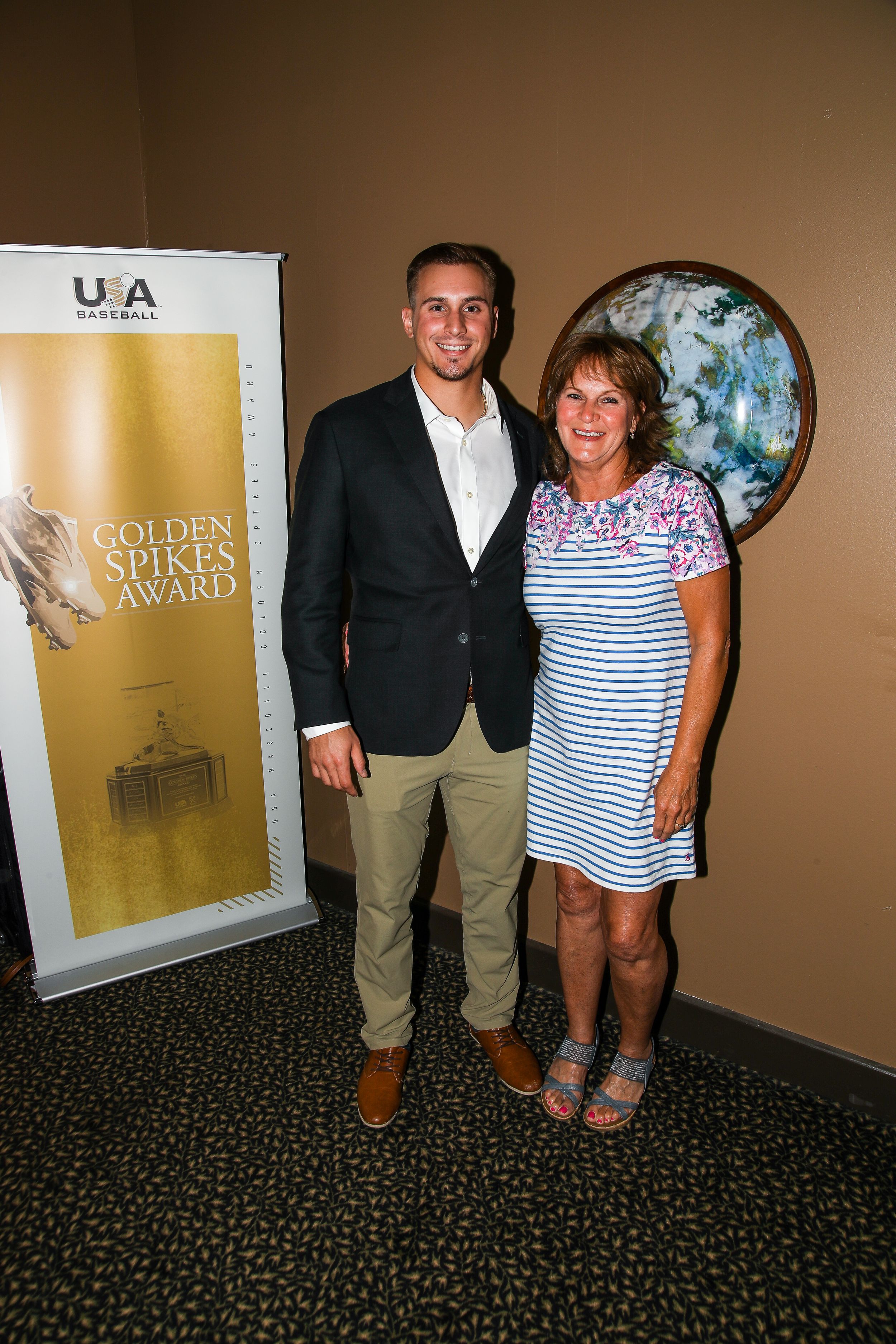 Man and woman posing together beside the USA Baseball Golden Spikes Award banner at an event.