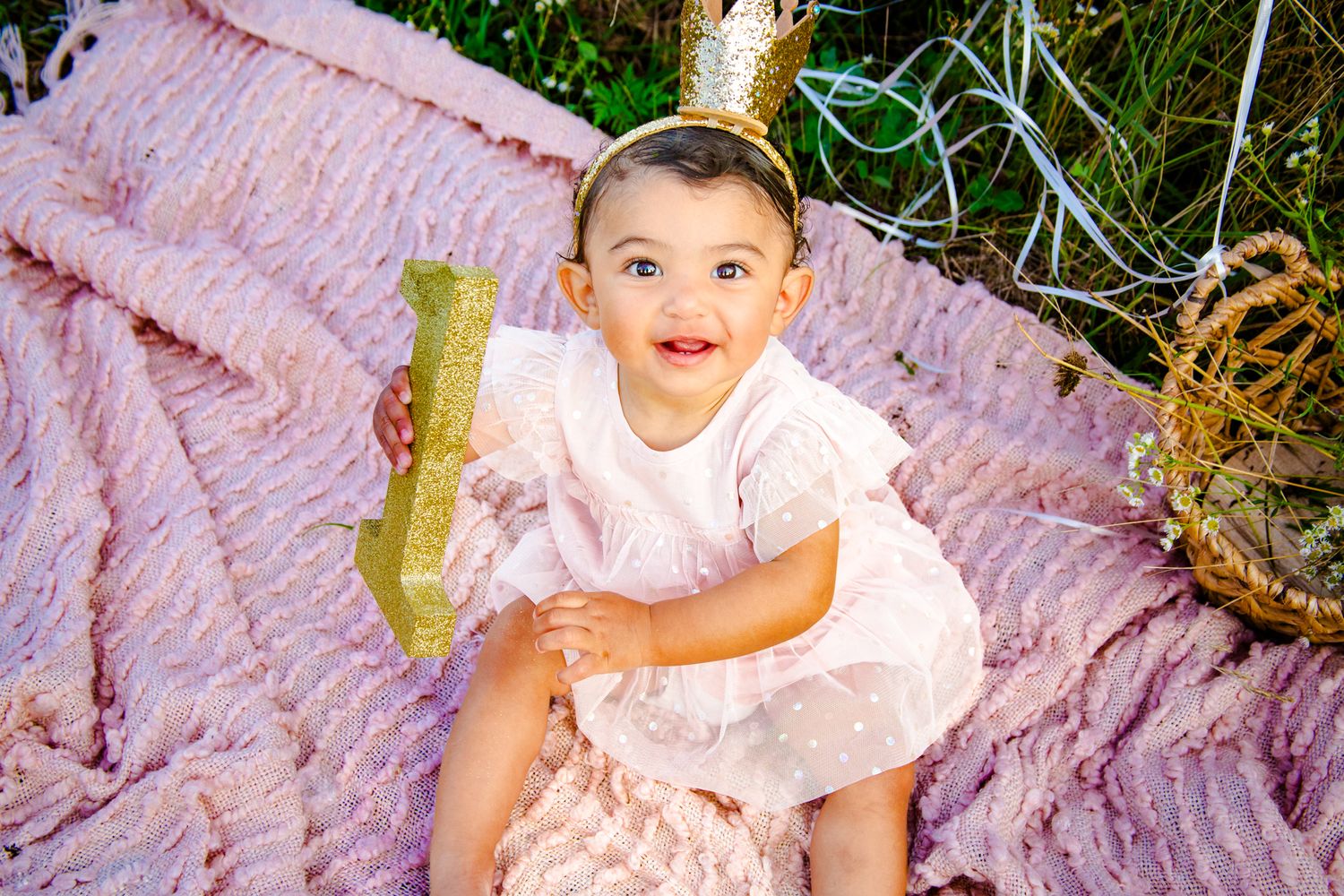 Baby in white dress sitting on pink blanket outdoors during summer.