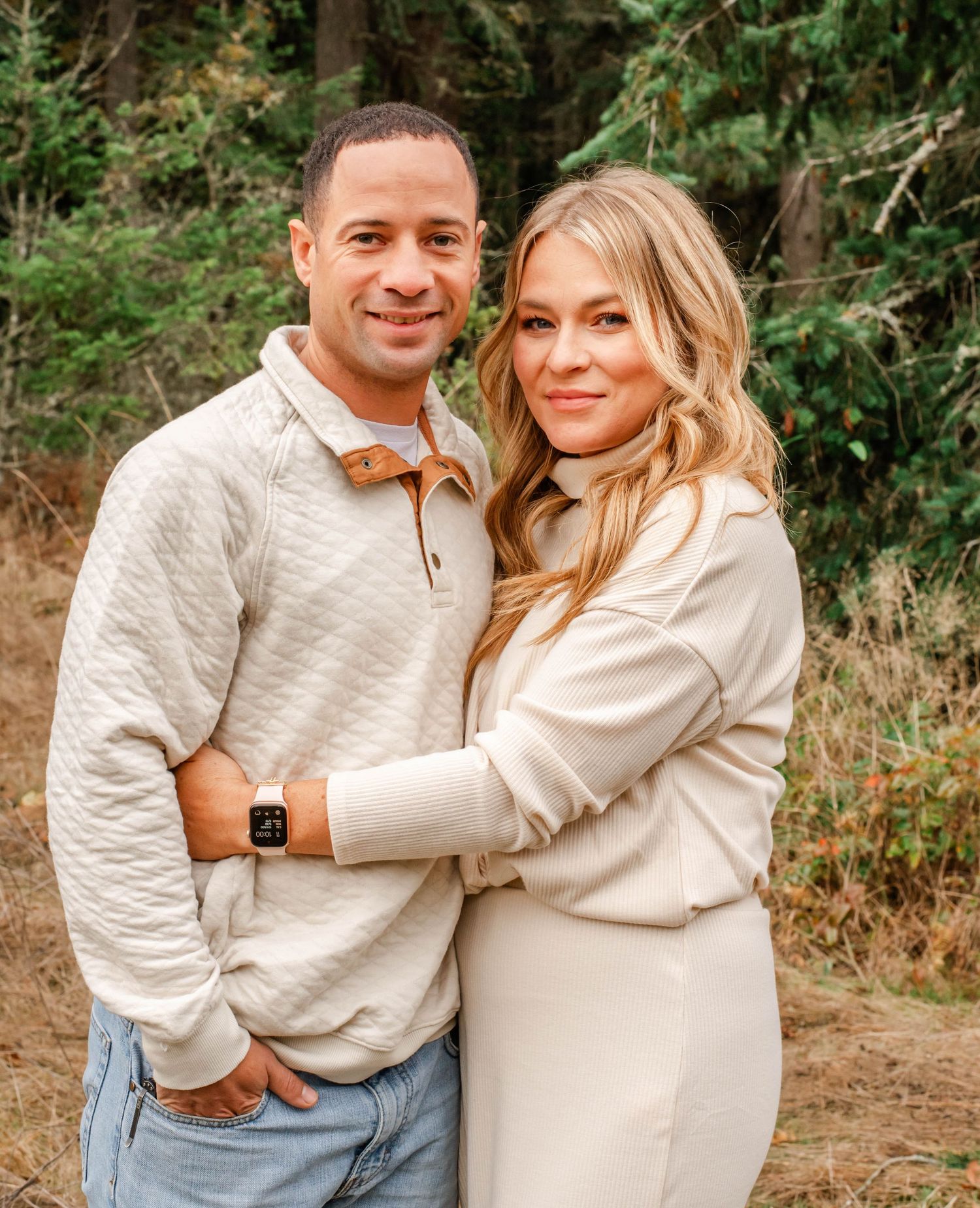 A couple in matching cream sweaters share intimate moments while posing together outdoors in a wooded setting.
