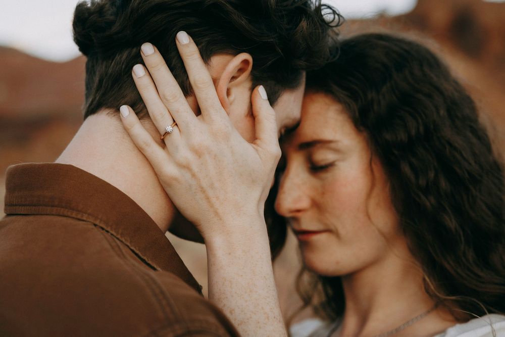 Couple sharing an intimate moment, foreheads touching, woman displaying engagement ring in desert setting.