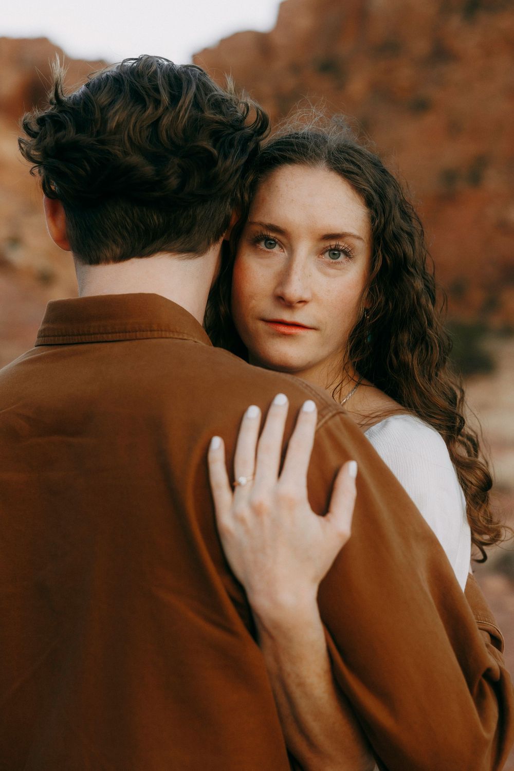 Woman with curly hair embracing man in brown jacket, showing engagement ring, red rock backdrop.