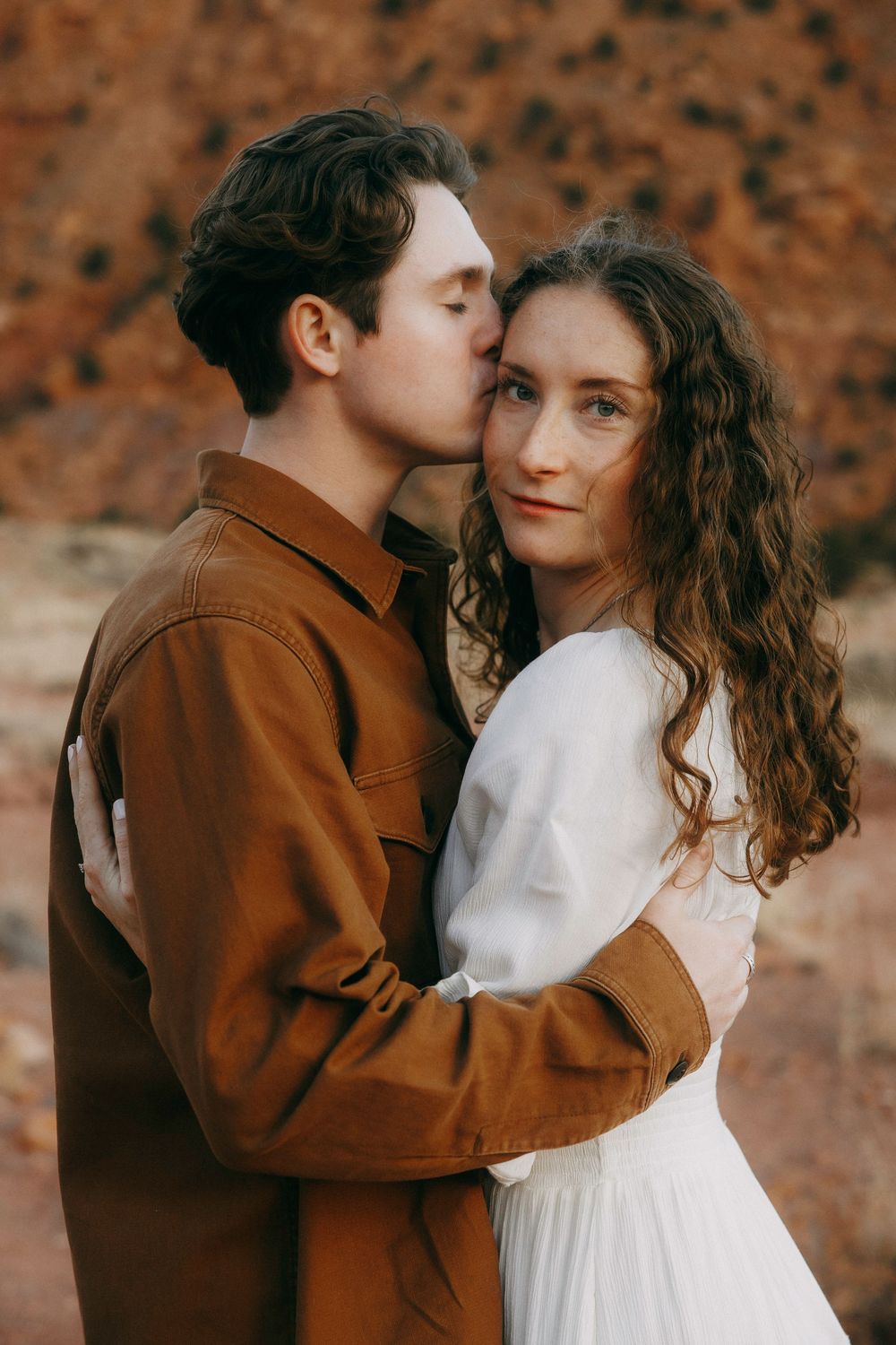 Man in brown jacket kissing woman's temple while embracing her in white dress outdoors.