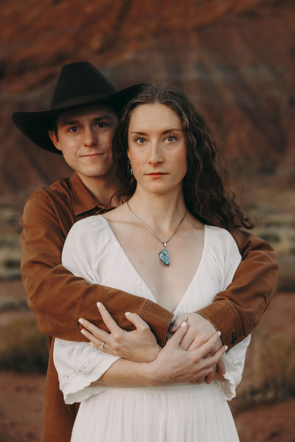 Couple posing in desert, man in cowboy hat and brown jacket embracing woman in white dress.