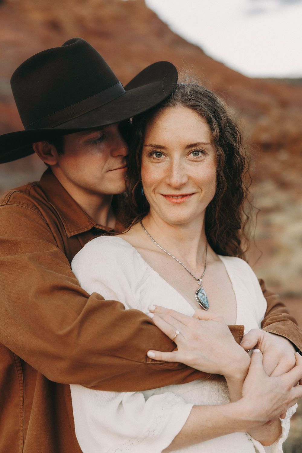 Smiling woman in white dress with turquoise necklace embraced by man in black cowboy hat.