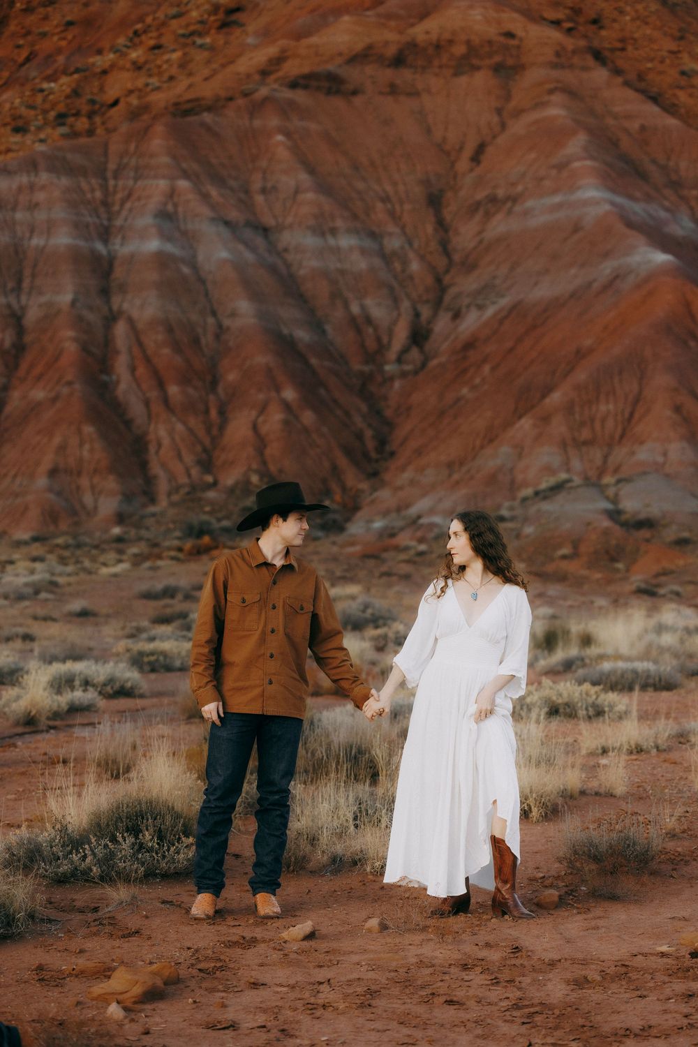 Engaged couple holding hands in red desert landscape, woman in white dress and cowboy boots.