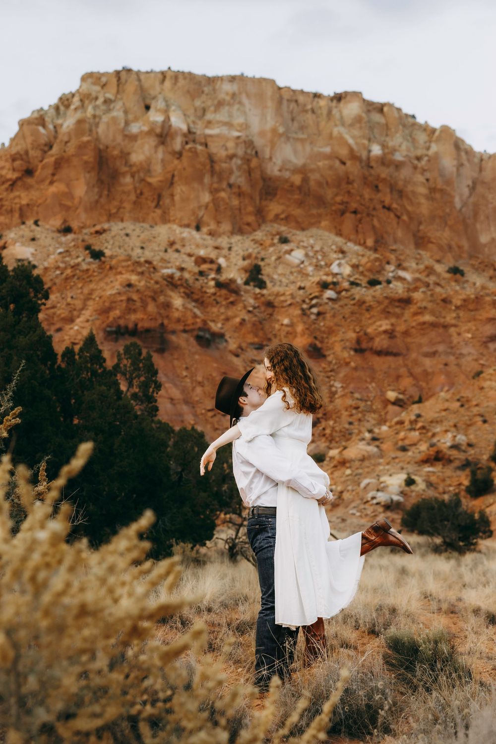 Man lifting woman in white dress in front of dramatic red rock canyon landscape.