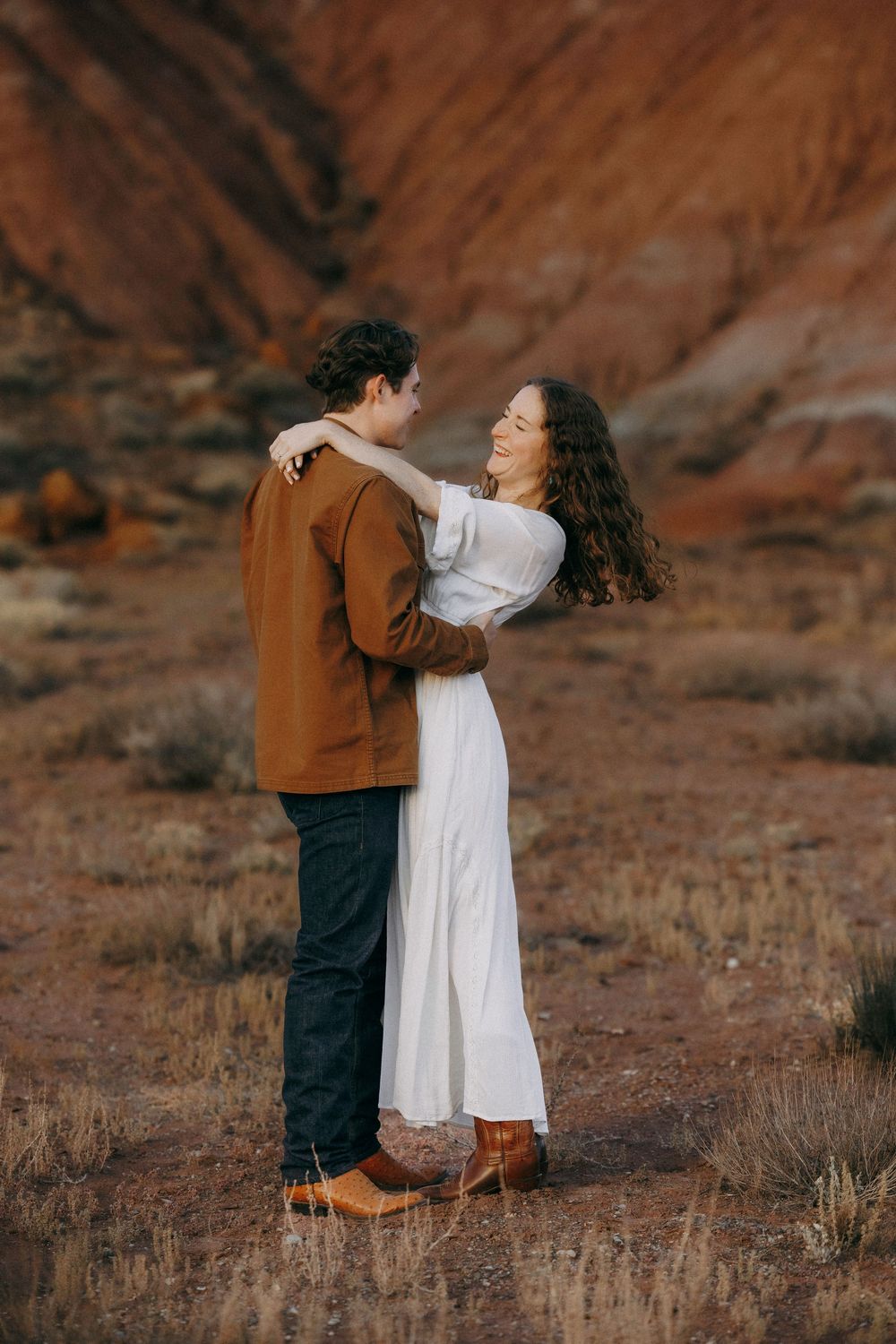 Couple dancing and embracing in white dress and brown jacket against red rock formations.