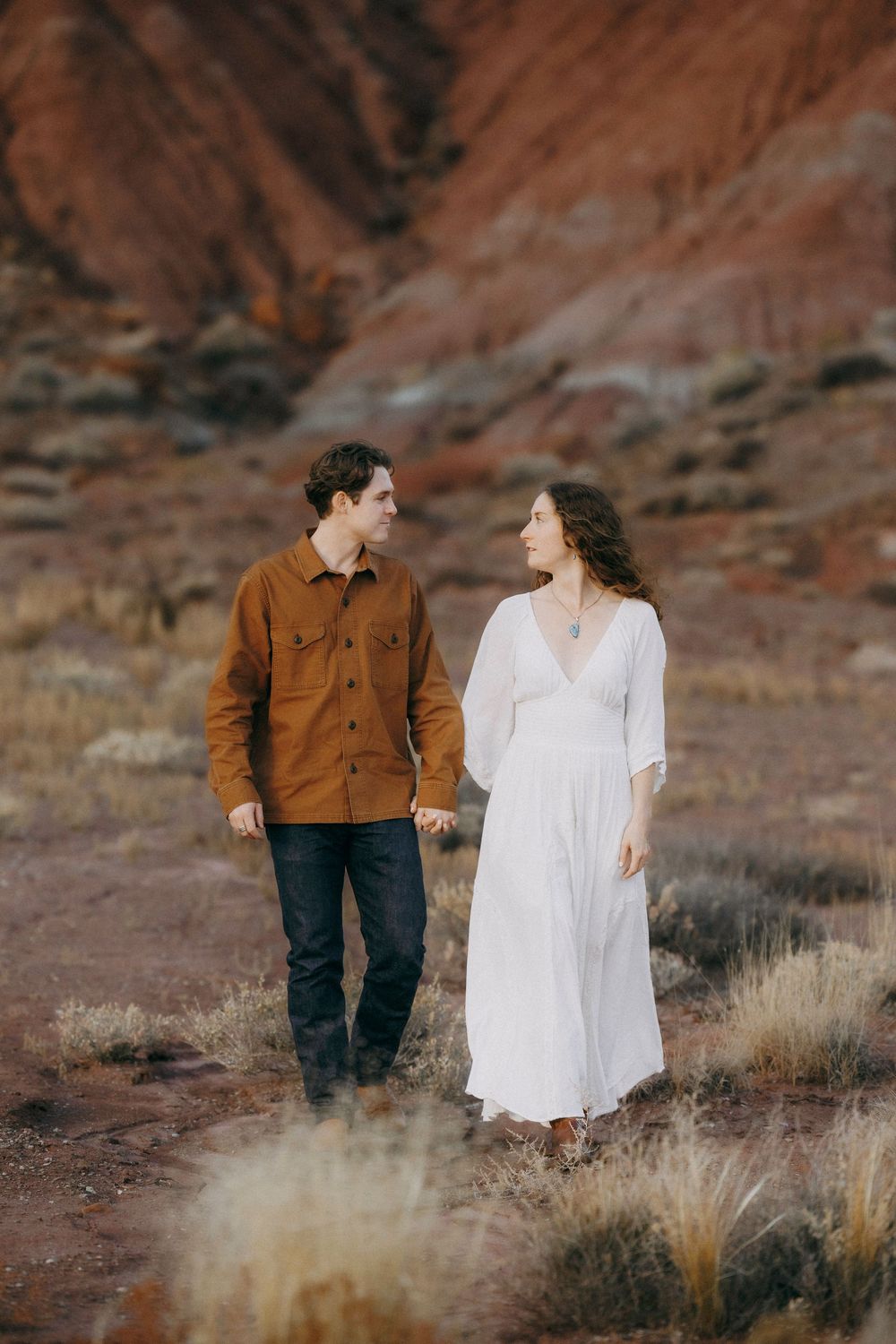 Couple walking hand in hand through desert landscape with vibrant red canyon backdrop.