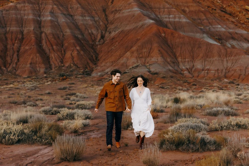 Couple running joyfully through open desert with dramatic red and orange canyon behind them.