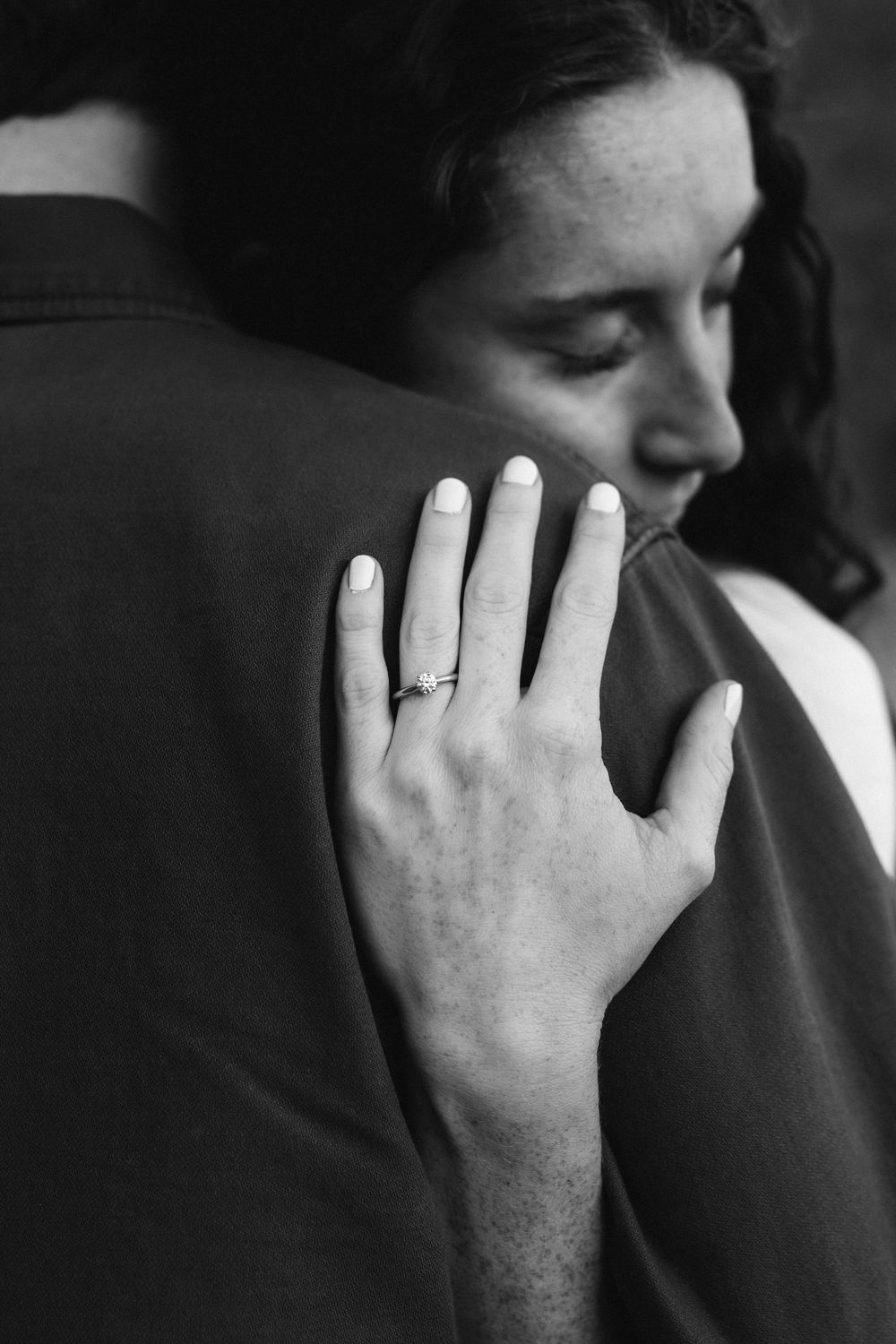 Close-up black and white photo of woman's hand with engagement ring resting on man's shoulder.
