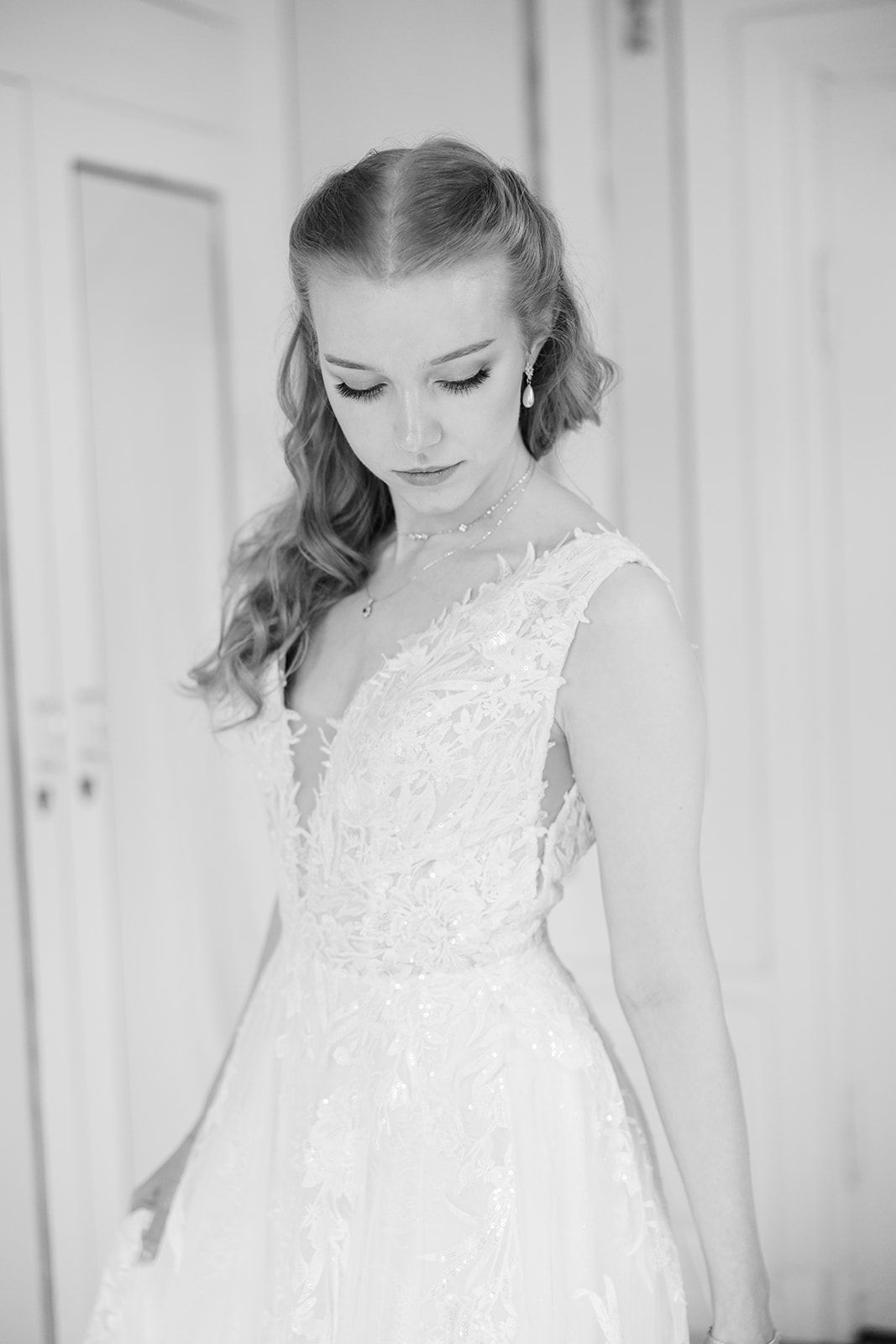A bride in a white lace wedding dress looks down gracefully in a bright, minimalist setting.