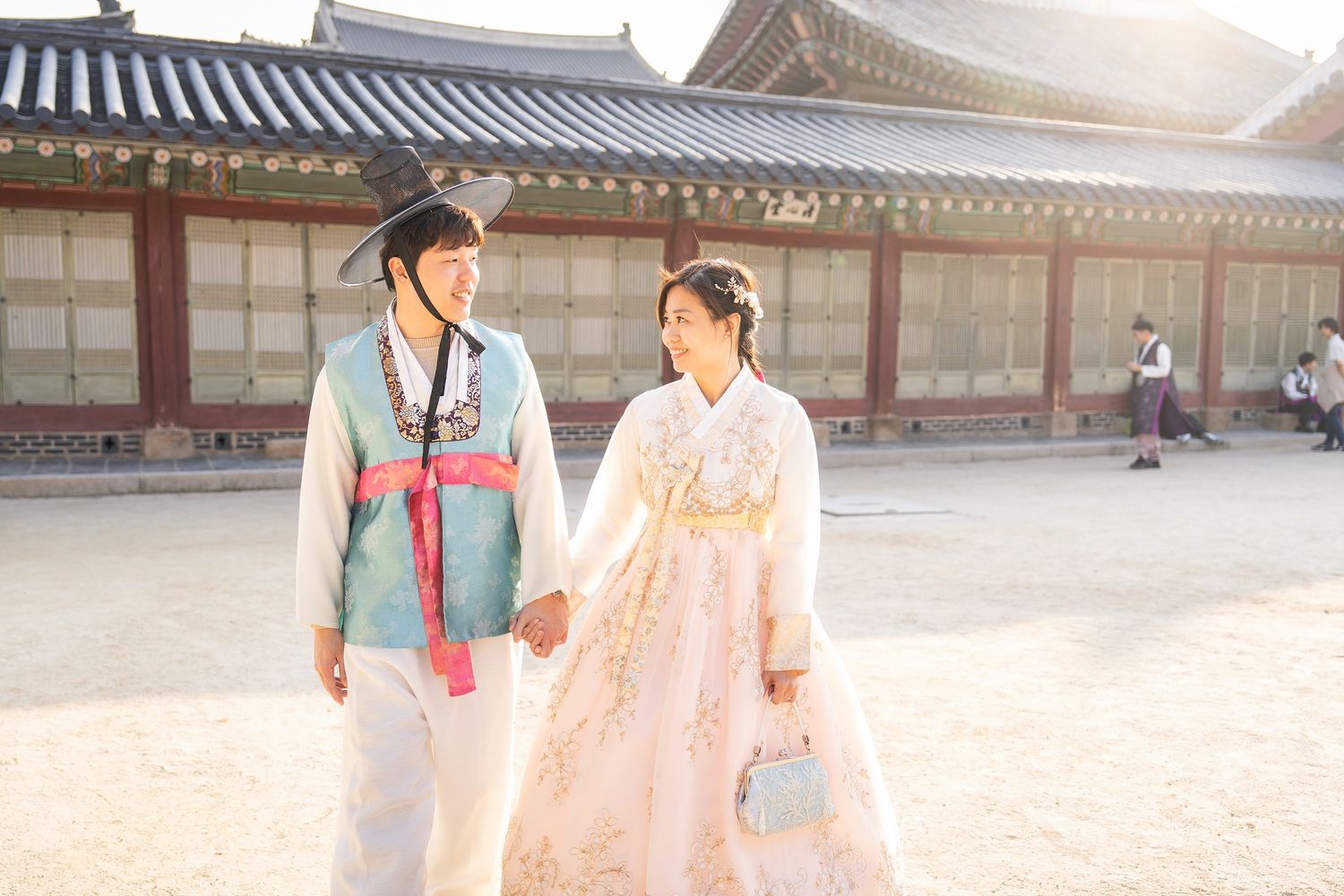 People wearing traditional Korean hanbok clothing walk through a historic palace courtyard on a sunny day.