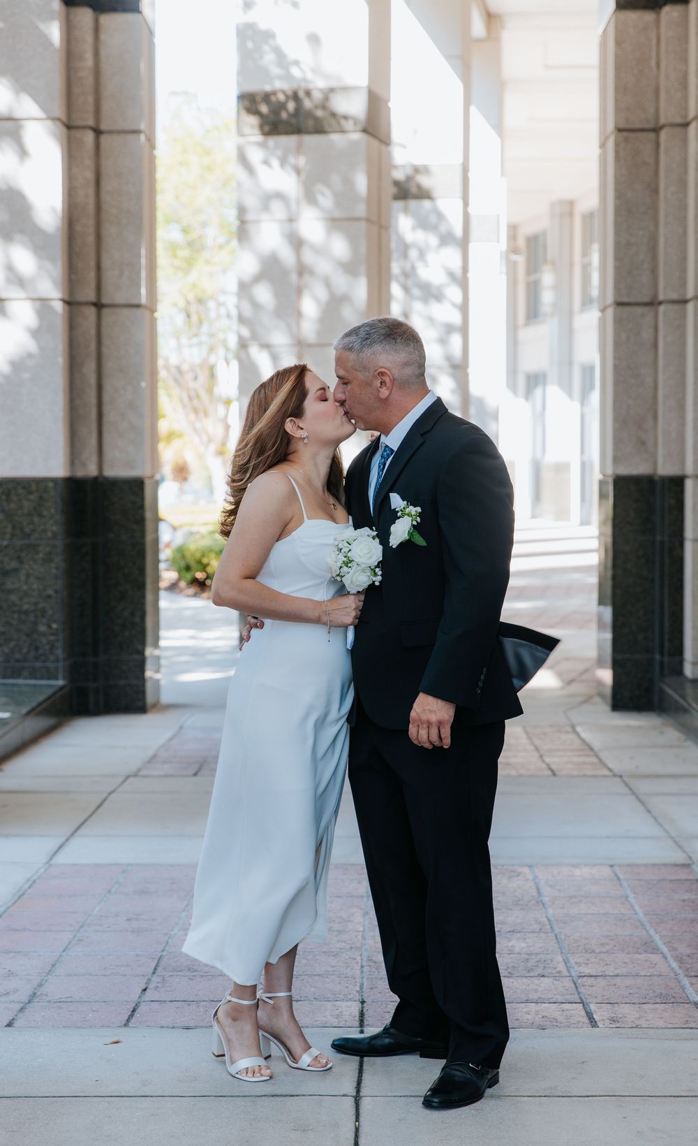 A romantic kiss between a couple in formal attire under columned architecture.