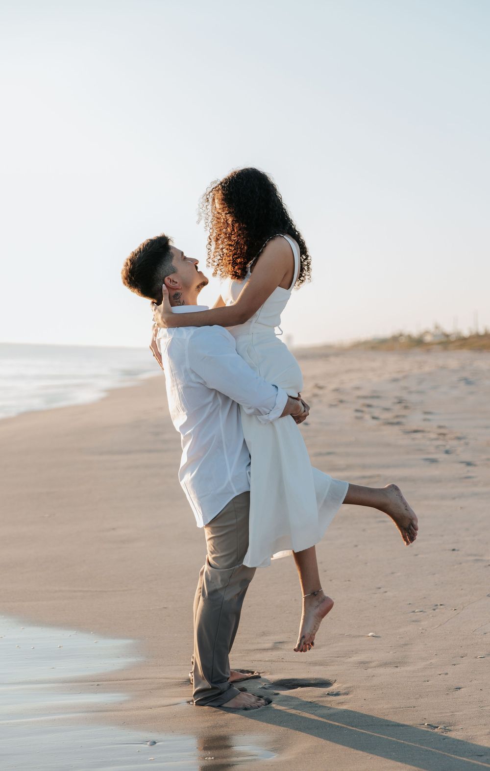 Joyful beach embrace with white attire against sunset backdrop.