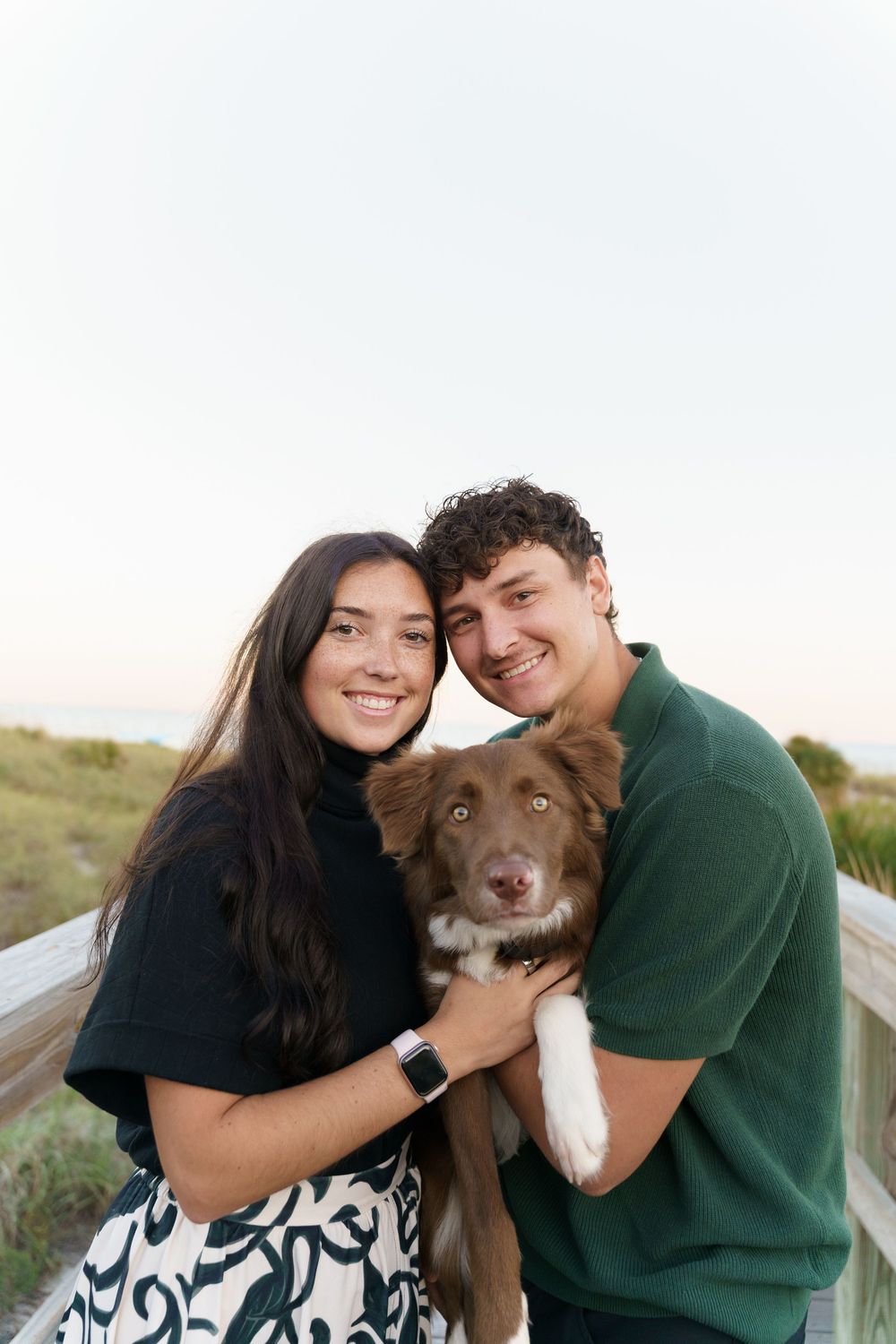 A couple poses outdoors with their brown and white dog in a series of affectionate photos at sunset.