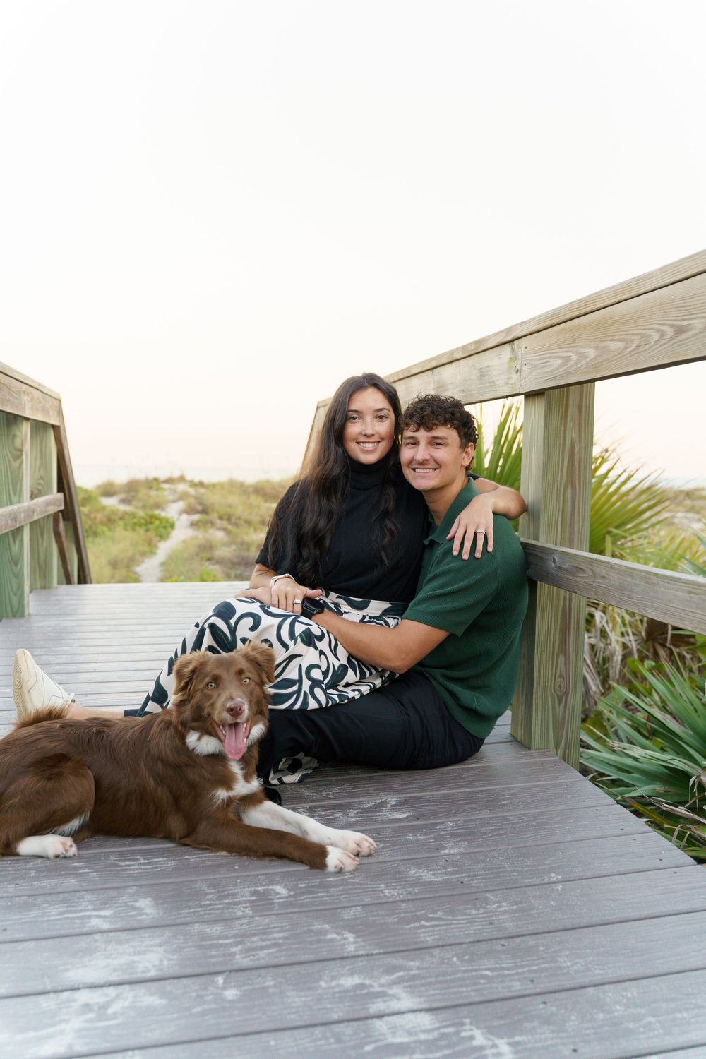 A couple and their brown dog relax together on a wooden deck surrounded by coastal plants.