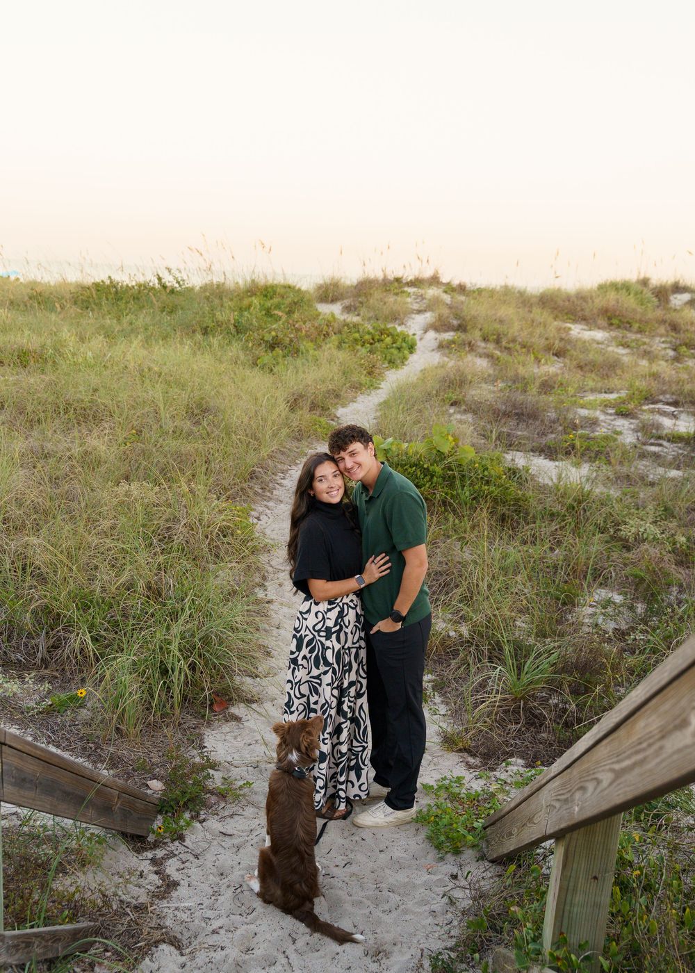 A couple walks along a sandy beach path through sea grass at sunset, sharing romantic moments together.