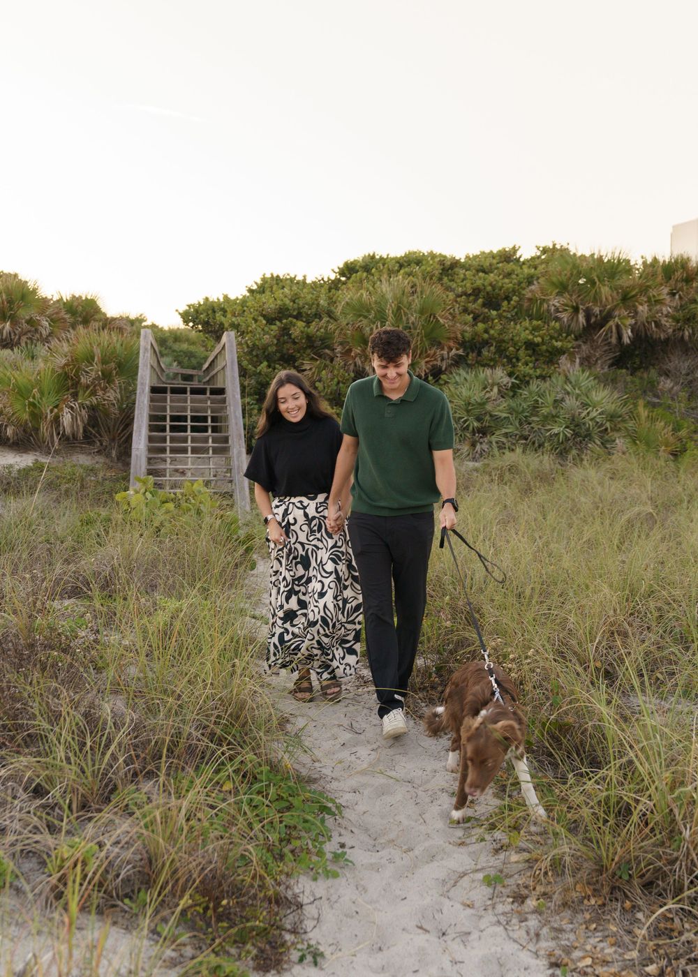 A couple walks their brown dog along a sandy beach path through coastal vegetation at sunset.