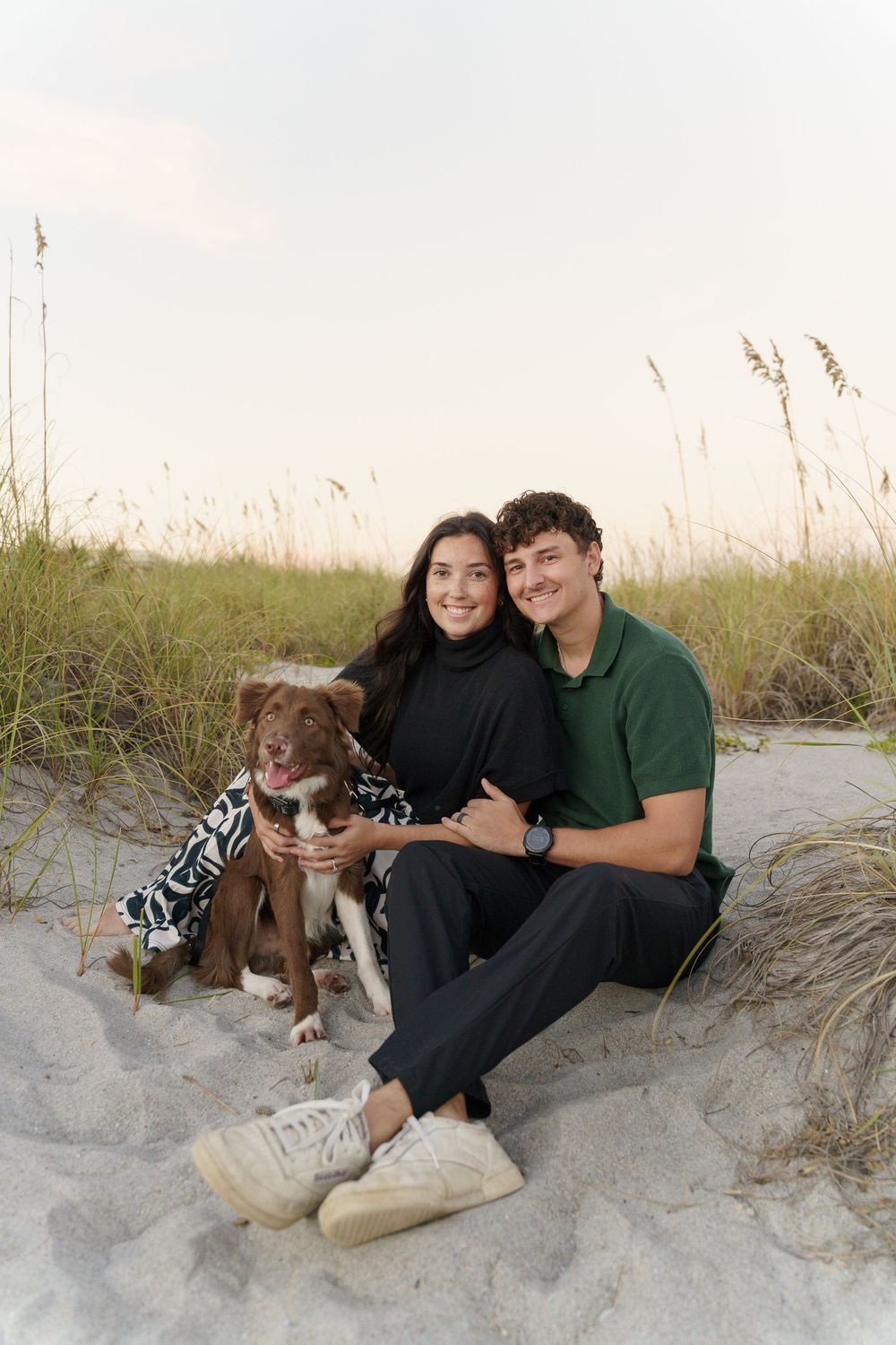 A couple sits with their dog on sandy dunes at the beach during golden hour.