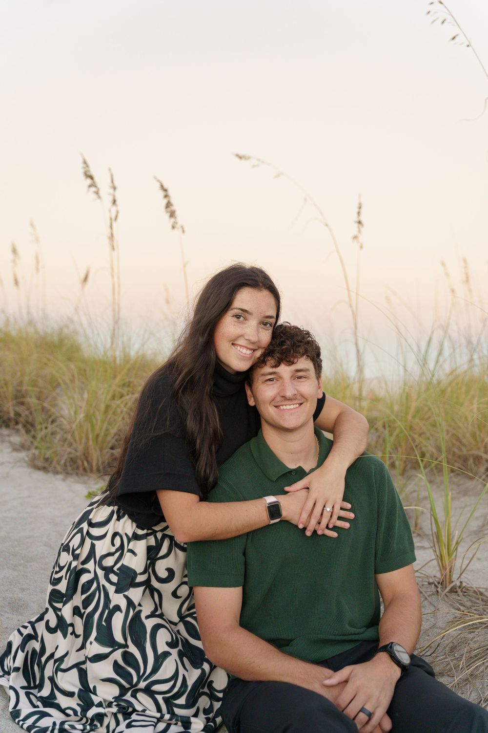 A couple sits together on beach dunes at sunset wearing green and patterned outfits.