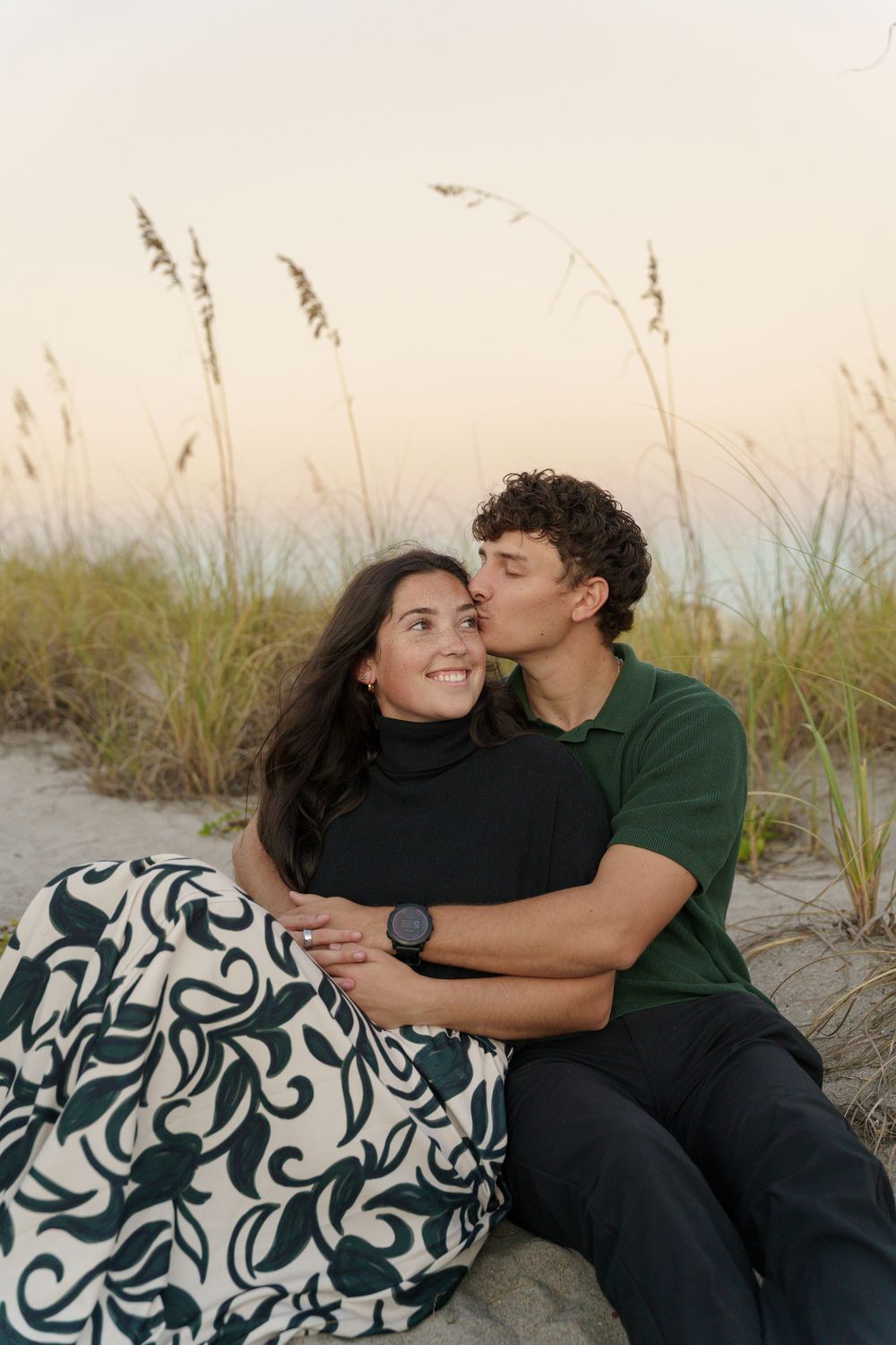 A couple shares tender moments on a sandy beach with sea grass at sunset.