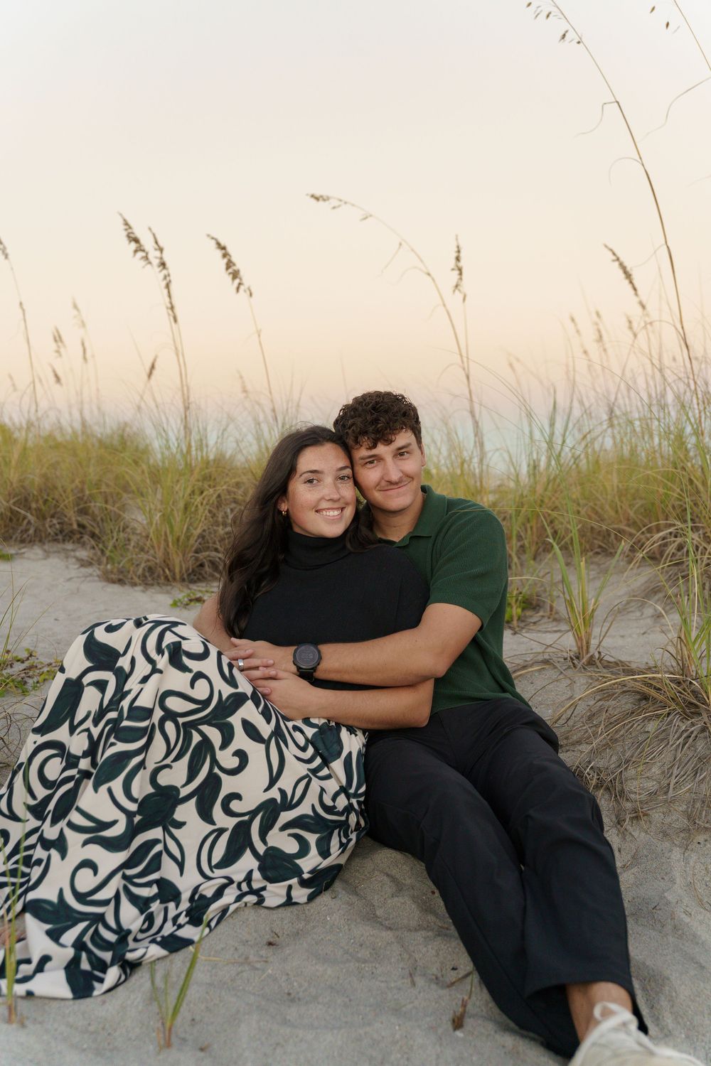 A couple embraces while sitting on a beach with coastal grass in the background.