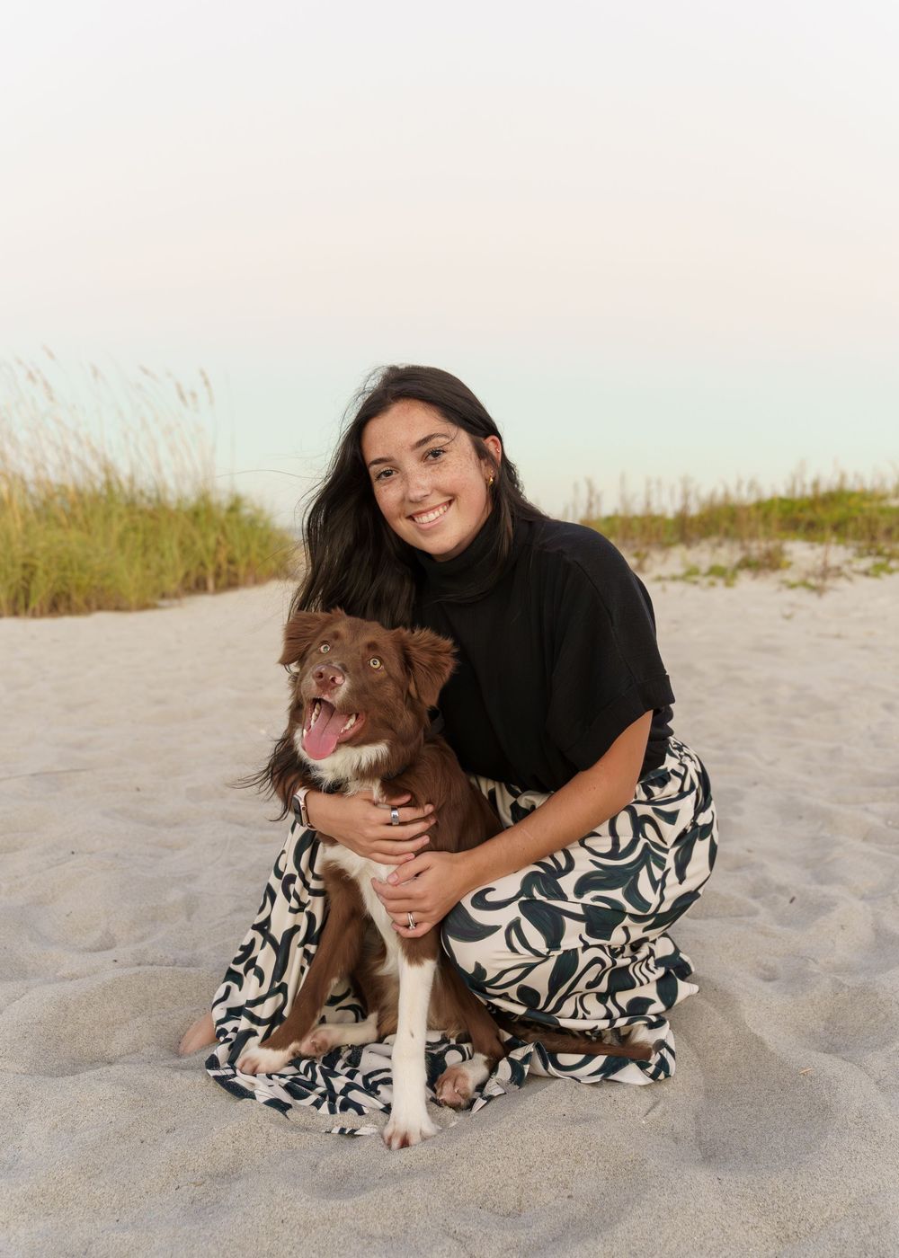A person in a black sweater and patterned skirt sits on a sandy beach with their brown and white dog.