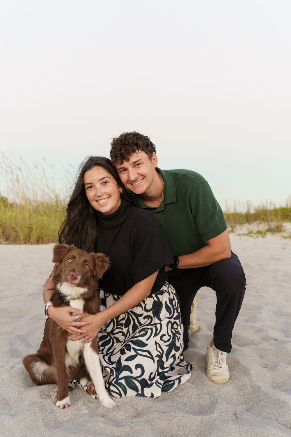 A couple poses with their brown and white dog on a sandy beach at dusk.