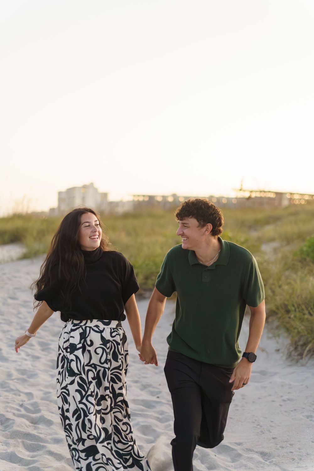 A couple in casual attire walks and laughs together along a sandy beach path at sunset.