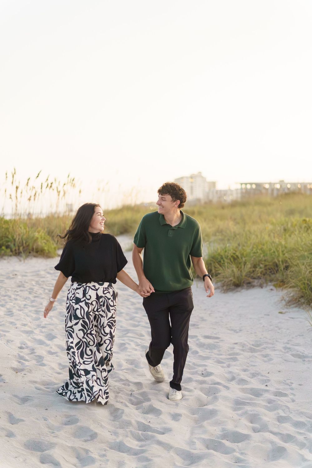 A couple walking hand in hand on a sandy beach path at sunset wearing coordinated black and green outfits.