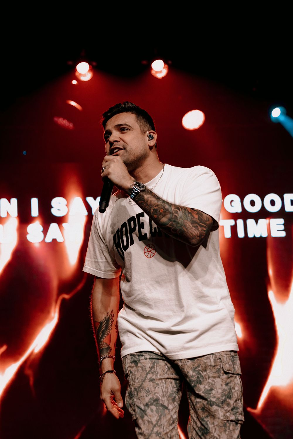 Concert performer in white t-shirt and camo pants sings on stage with dramatic red lighting and illuminated text display.